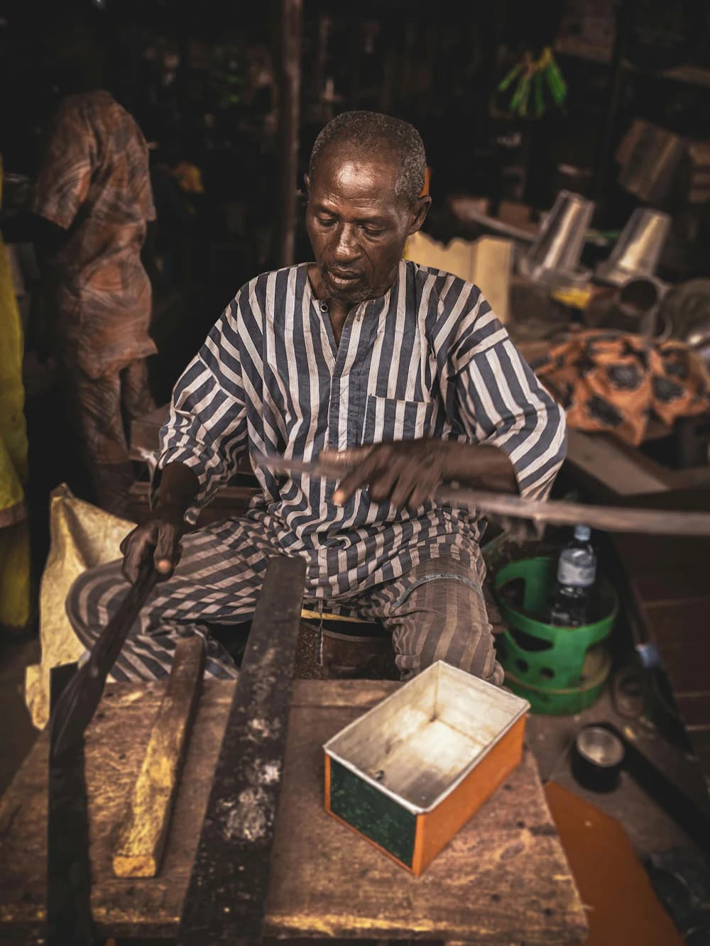 An older craftsman in a striped shirt shaping wood at his workbench.