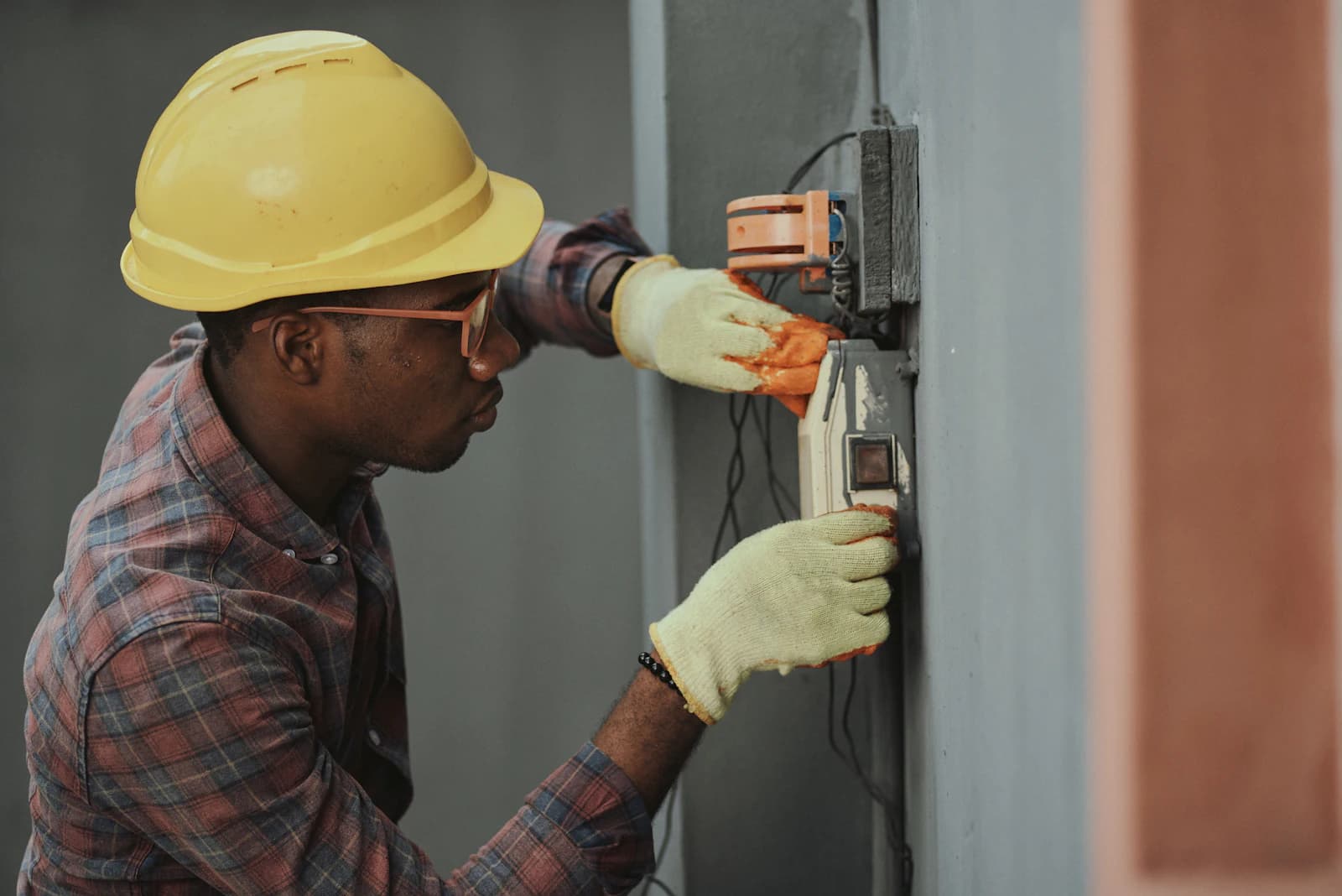 An electrician in a hard hat and gloves repairing a wall socket.